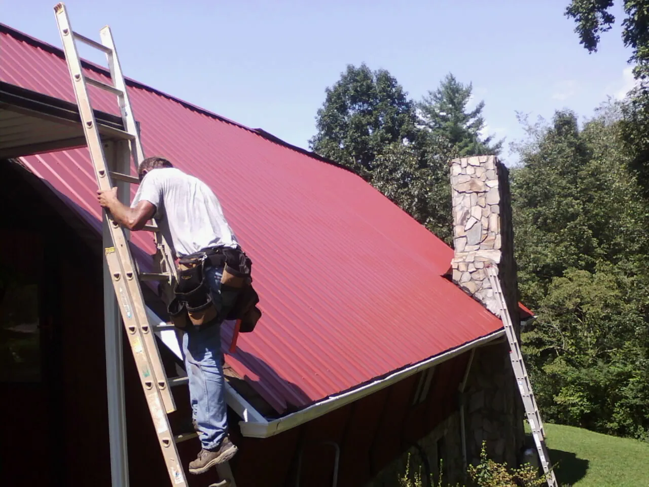Roofer on a ladder beside a red metal roof and stone chimney — Soffit & Fascia Repair by Roofing Brothers in Bruceton